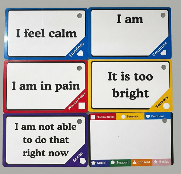 A photograph showing six color-coded communication cards arranged in two columns on a neutral grey background. The top-left blue card reads "I feel calm" (Emotions). Beside it, another blue card reads "I am" followed by blank space. The middle-left red card says "I am in pain" (Physical Needs). The middle-right yellow card says "It is too bright" (Sensory). The bottom-left purple card reads "I am not able to do that right now" (Social). The bottom-right is a blank index card showing color category keys.